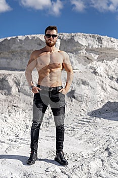 Halfnaked strong man in sunglasses standing in beautiful location. Bodybuilder posing