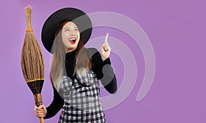 Halloweeen theme, young pretty asian woman in witch costume holding broom posing finger pointing on purple background