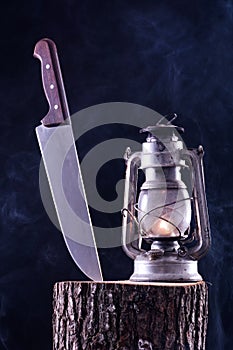 Halloween background of big knife stabbed in wood log and old burning gas lantern