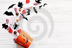 Halloween candy with skulls, black bats, ghost, spider decorations spilled from jack o lantern bucket on white wooden background,