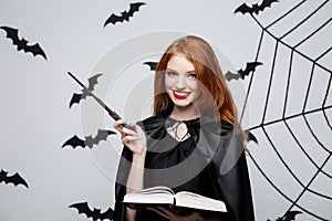 Halloween Concept - Beautiful Witch playing with magic stick and magic book on grey background.