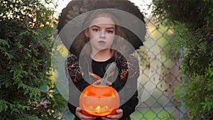 Halloween. Cute girl shows a cheerful witch. She is dressed in a black dress and hat. Girl holding pumpkin with a
