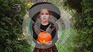 Halloween. Cute girl shows a cheerful witch. She is dressed in a black dress and hat. In the hands of the girl are