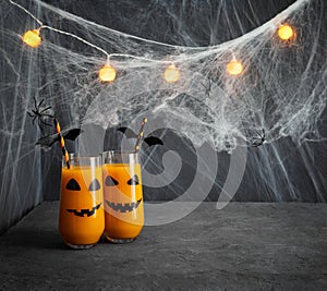 Halloween drink on a dark gray background with glowing garlands. Pumpkin or carrot juice in glasses with jack-o-lantern face and