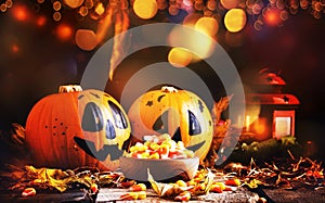 Halloween festive composition with sweet corn in bowl and smiling pumpkins guards, lantern, straw and fallen leaves on dark wooden