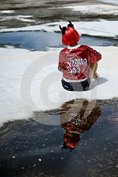 Halloween. The girl in the red wig with horns and a red jacket sitting near the puddle with his reflection. back to the