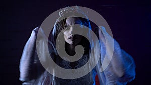 Halloween girl with scary make-up and wreath of dry flowers on head standing moving hands in veil looking at the camera