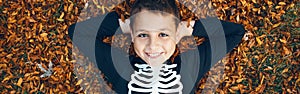 Halloween holiday. Happy child boy in skeleton costume lying on pile of yellow leaves outdoors. Smiling boy preparing for