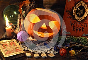 Halloween Jack O Lantern Pumpkin with grimoire book, tarot card Devil and runes on witch table