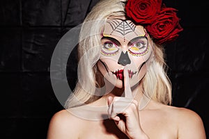 Halloween Make-Up Style. Blond Model Wear Sugar Skull Makeup with Red Roses. Santa Muerte Concept. Silence Gesture