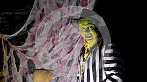 Halloween party, night, twilight, in the rays of light, a man with a terrible make-up, with a green face and a hat shows