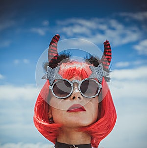 Halloween. Portrait of a beautiful girl in a red wig with horns close-up against a blue sky. Red, holiday makeup.