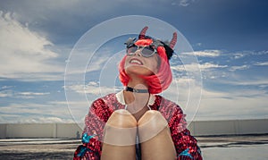 Halloween. Portrait of a beautiful girl in a red wig with horns close-up against a blue sky. Sitting on the foof. Red