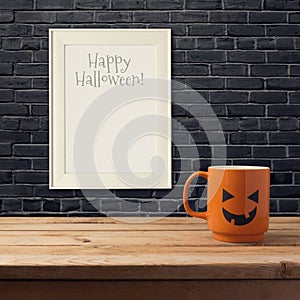 Halloween poster mock up template. Coffee cup as jack o lantern pumpkin on wooden table over black brick wall.