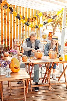 Halloween Preparaton Concept. Young couple sitting at table outdoors making jack-o`-lantern carving pumpkins joyful