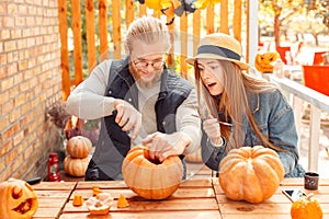 Halloween Preparaton Concept. Young couple sitting at table outdoors making jack-o`-lantern guy carving pumpking while