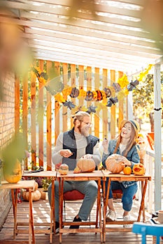 Halloween Preparaton Concept. Young couple sitting at table outdoors making jack-o`-lantern laughing playful