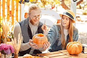 Halloween Preparaton Concept. Young couple sitting at table outdoors making jack-o`-lantern painting face on pumpkin