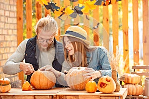 Halloween Preparaton Concept. Young couple sitting at table outdoors making jack-o`-lantern smiling cheerful