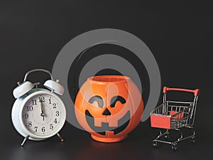 Halloween pumpkin bucket  with shopping cart and white vintage alarm clock 12 pm. isolated  on black  background with copy space.