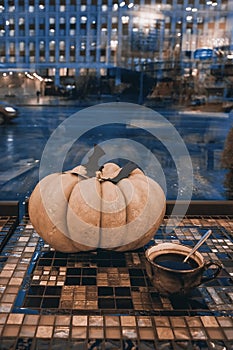 Halloween pumpkin with carved black bats and cup of coffee on table by the window, dark city dusk, traditional decor