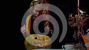 Halloween pumpkin with light inside on table in night forest with blurred little girl opening vegetable warming hands in