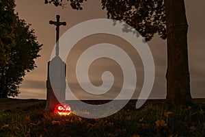 Halloween pumpkin beside a way cross with leaves and tree trunk