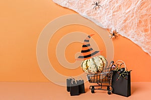 Halloween shopping holiday concept, pumpkin in grocery cart with black paper bags on orange background with spider web