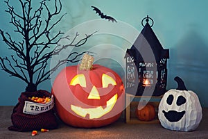 Halloween still life scene with carved pumpkin, lantern, treats bag, and ghost gourd with teal background with night shadows.  It