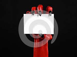 Halloween theme: Red devil hand with black nails holding a blank white card on a black background