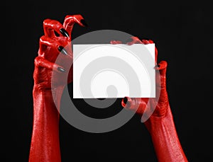 Halloween theme: Red devil hand with black nails holding a blank white card on a black background