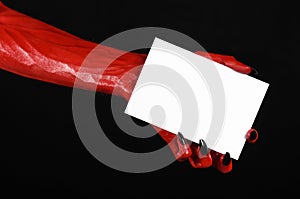 Halloween theme: Red devil hand with black nails holding a blank white card on a black background