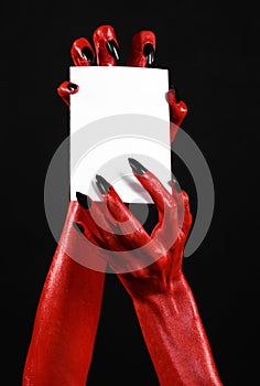 Halloween theme: Red devil hand with black nails holding a blank white card on a black background