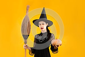 Halloween theme, young asian woman in black dress witch costume wearing black witch hat holding broom and pumpkin posing on yellow