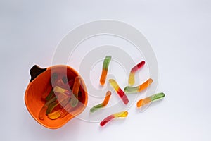 Halloween treats gummy worms in pumpkin bowl. Colorful jelly candies on white background. Top view