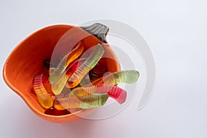 Halloween treats gummy worms in pumpkin bowl. Colorful jelly candies on white background. Top view