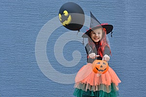 Halloween trick or treat. Portrait smiling girl in witch hat with balloon and pumpkin bucket for sweets against a blue wall . Copy