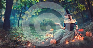 Halloween Witch with a carved pumpkin and magic lights in a dark forest at night. Beautiful young woman in witches costume photo