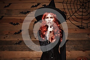 Halloween witch concept - Happy Halloween red hair Witch doing silence gesture with finger on her lips over old wooden