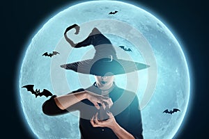 Halloween witch girl conjures against the background of the moon. Beautiful young woman in a witch hat. Halloween parties, copy photo