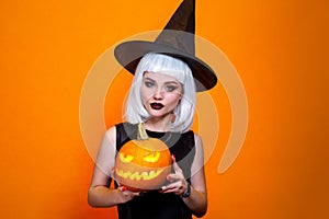 Halloween witch in hat and costume holding carved pumpkin on orange background.