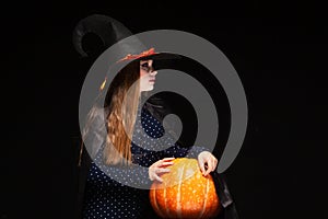 Halloween Witch with Pumpkin on black background. Beautiful young surprised woman in witches hat and costume holding