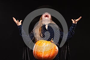 Halloween Witch with Pumpkin on black background laughs evilly. Beautiful young surprised woman in witches hat and