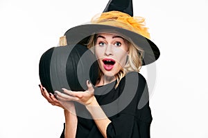 Halloween Witch with shocked expression holding large black pumpkin. Beautiful young woman in witch hat holding pumpkin.