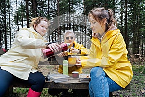 Halt for snack during hiking. Company hikers enjoying picnic on the bench, drinking hot tea, eating sandwiches in forest