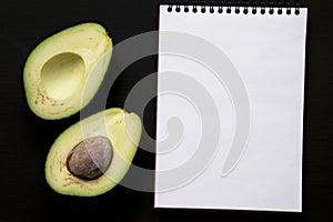 Halved avocado and blank notepad over black background, top view. From above, overhead. Copy space