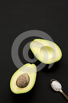 Halved avocado and sea salt over black surface, overhead view. Top view, from above.
