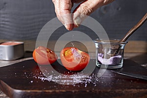 Halved tomato being sprinkled with salt on a wood cutting board
