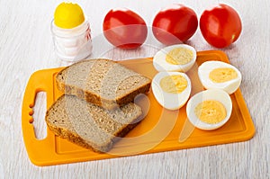 Halves of boiled eggs, bread on cutting board, salt shaker, tomatoes on wooden table