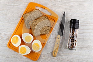 Halves of boiled eggs, bread on cutting board, knife, jar with condiment on wooden table. Top view